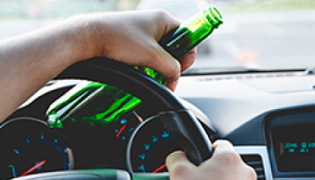 A person holding a steering wheel with one hand and a green bottle with the other hand while driving, indicating irresponsible drinking and driving behavior.